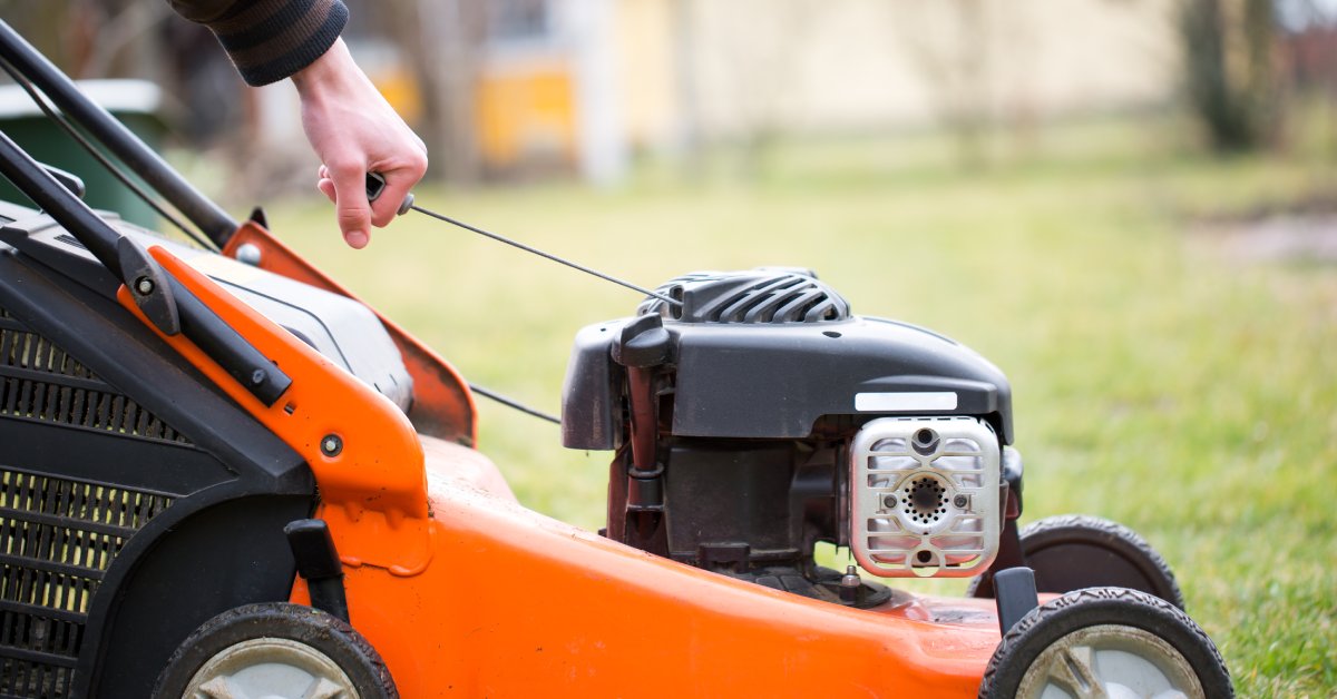 A person who's wearing a sweater holding the pull cord of a lawn mower. They are pulling it to start the engine.