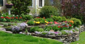 A layered garden with various types of flowers in front of a house. Each layer is separated by a wall of stones.