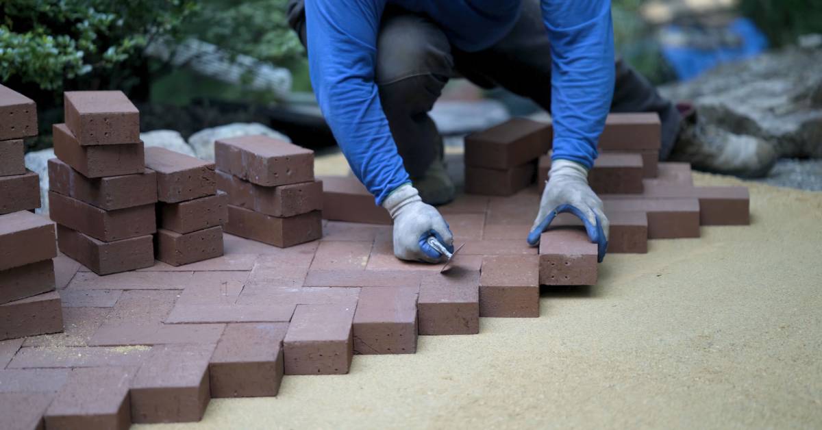 A person laying bricks for a backyard patio. The bricks are reddish-brown and placed diagonally in a zigzag pattern.