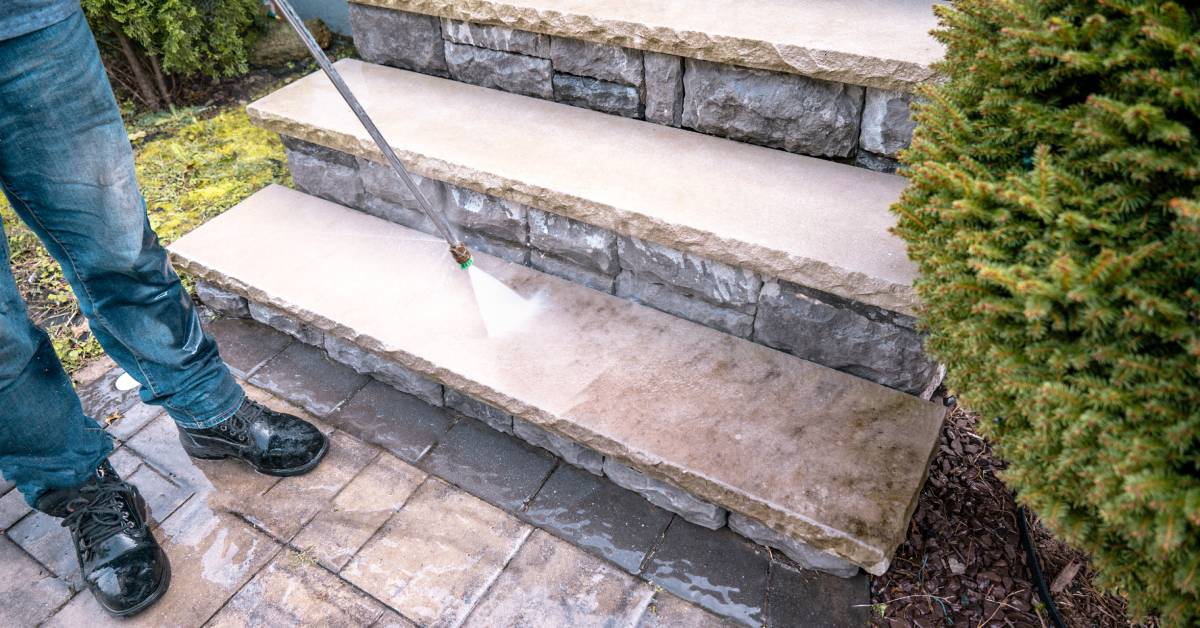 A man washing an outdoor set of stone steps with a power washer. There is a round bush next to the steps.