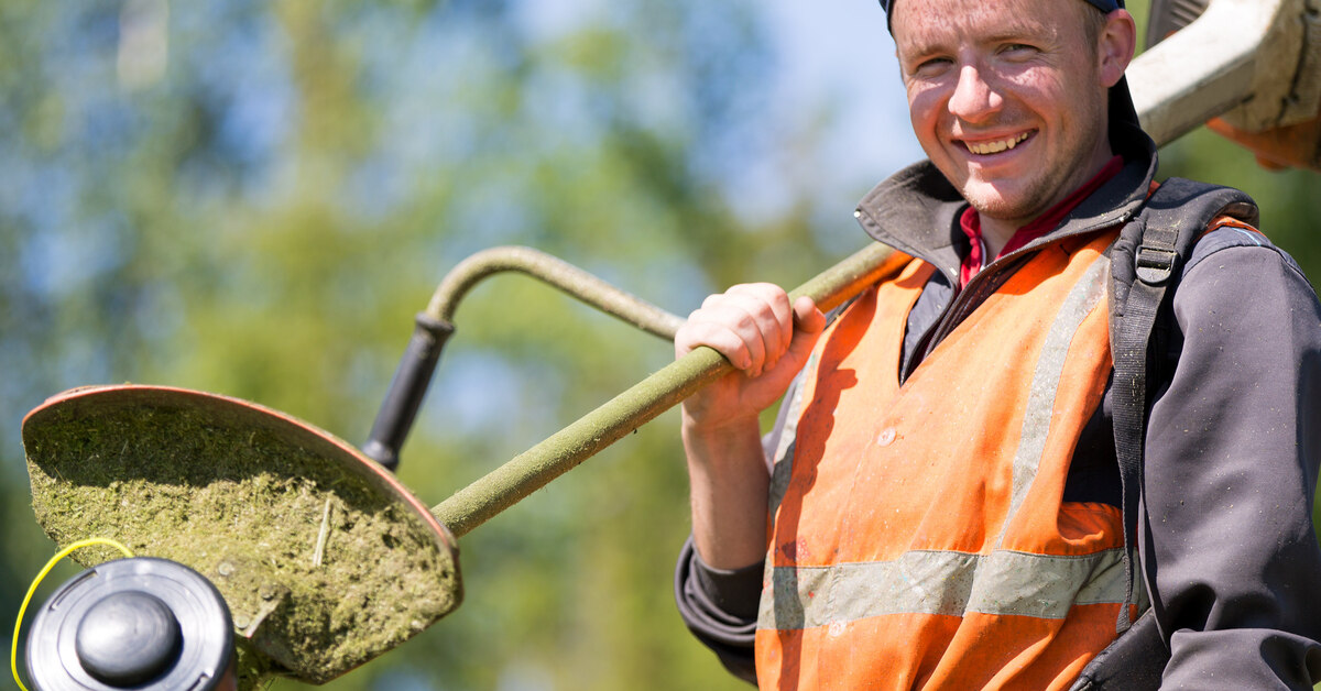 A landscaper holding a weed whacker over his shoulder. He is wearing a reflective vest and is smiling at the camera.