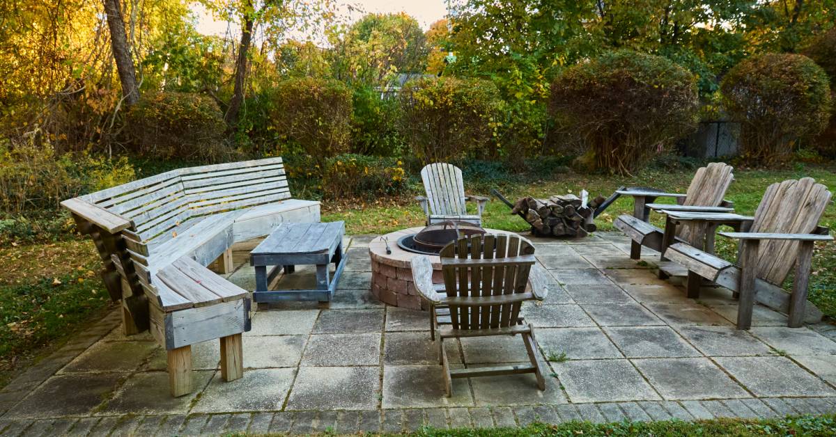 A stone fire pit surrounded by wooden chairs and benches. There is a pile of logs off to the side of the seating area. 