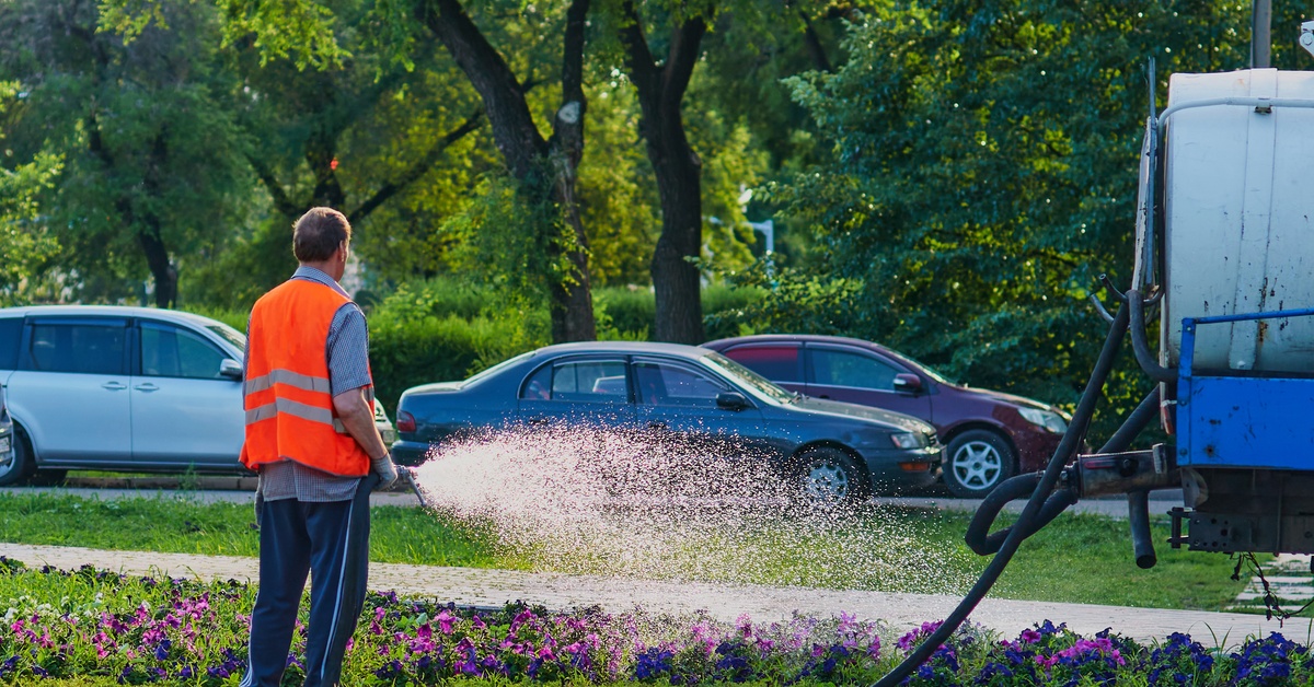 A worker in an orange vest sprays fine mist over a bed of purple petunias in a park setting.