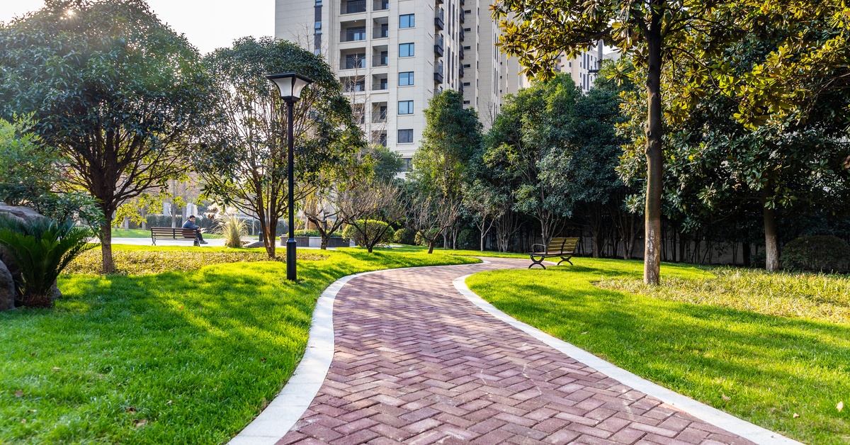 A curved brick pathway running through a landscaped park lawn bordered by benches and lamp posts.