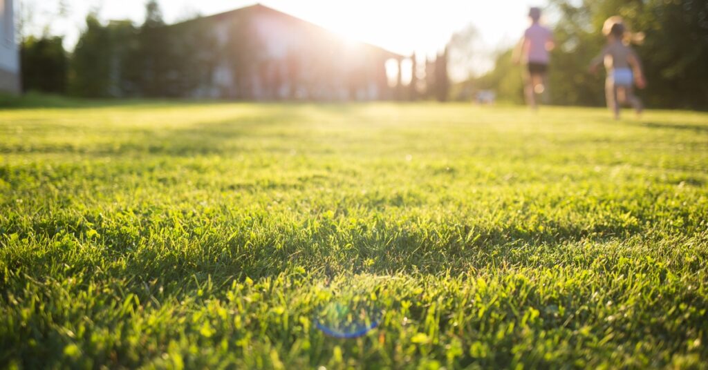 A brightly lit lawn with the sun setting behind a home in the background and children running out of focus.