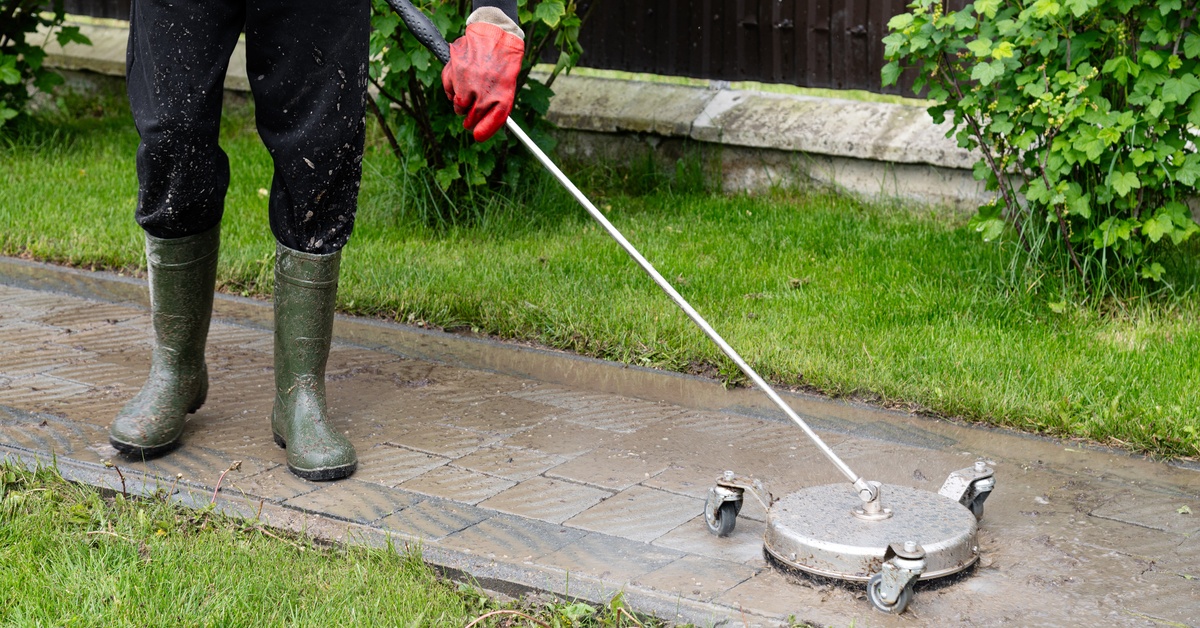 A person wearing waterproof boots, a long sleeve shirt, and red gloves pressure washing an outdoor stone walkway.