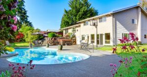 A large backyard with a curving pool, lawn chairs, hot tub, and garden flowers beside a beige two-story house.