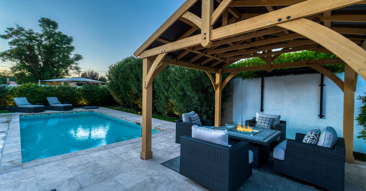 A square backyard pool beside a pergola with wicker furniture, a fire table, light blue rug, and light grey tile flooring.
