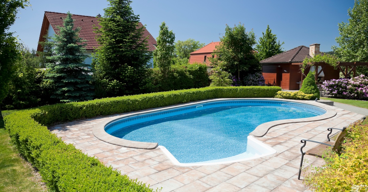 A backyard pool surrounded by stone pavers, hedges, patio chairs, and several houses with red roofs and green trees.