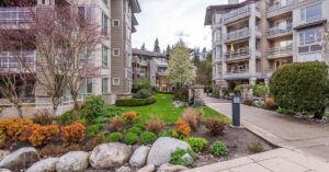 Pink flowers, green foliage, and black lantern-style lights along a concrete sidewalk by a residential building.