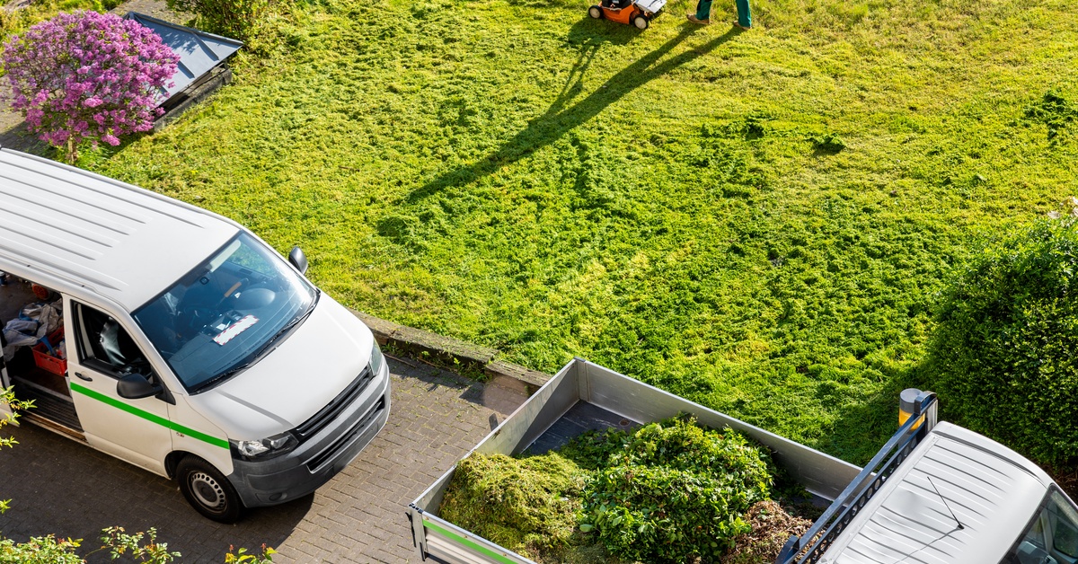 Aerial view of a worker mowing grass near two white utility vans and a trailer filled with green yard waste.