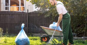 A man in green overalls uses a rake next to a wheelbarrow and full trash bags on a grassy backyard lawn.