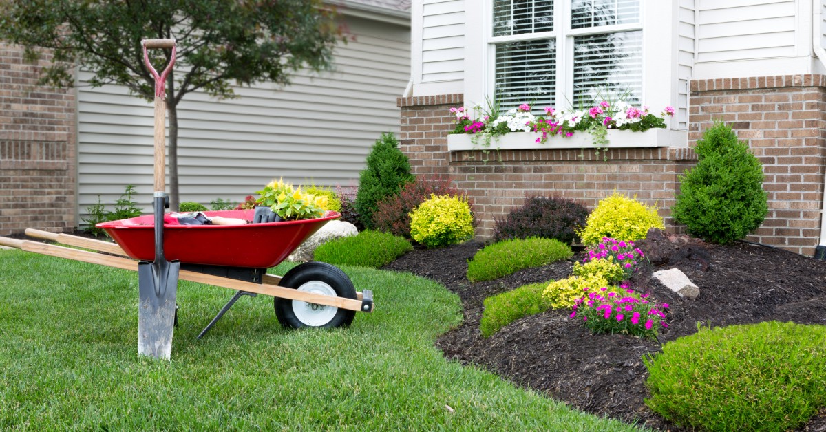 A shovel is leaning against a red wheelbarrow. There is a house in the background and the wheelbarrow is on grass.