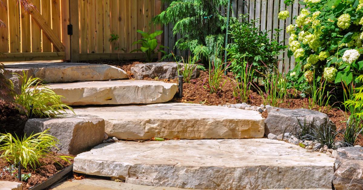 A garden pathway, bordered by plants, rocks, and shrubs, with large stone steps leading up to a wooden gate.