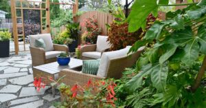 A patio seating area with wicker chairs, cushions, table, umbrella, and surrounding plants on a stone-paved surface.