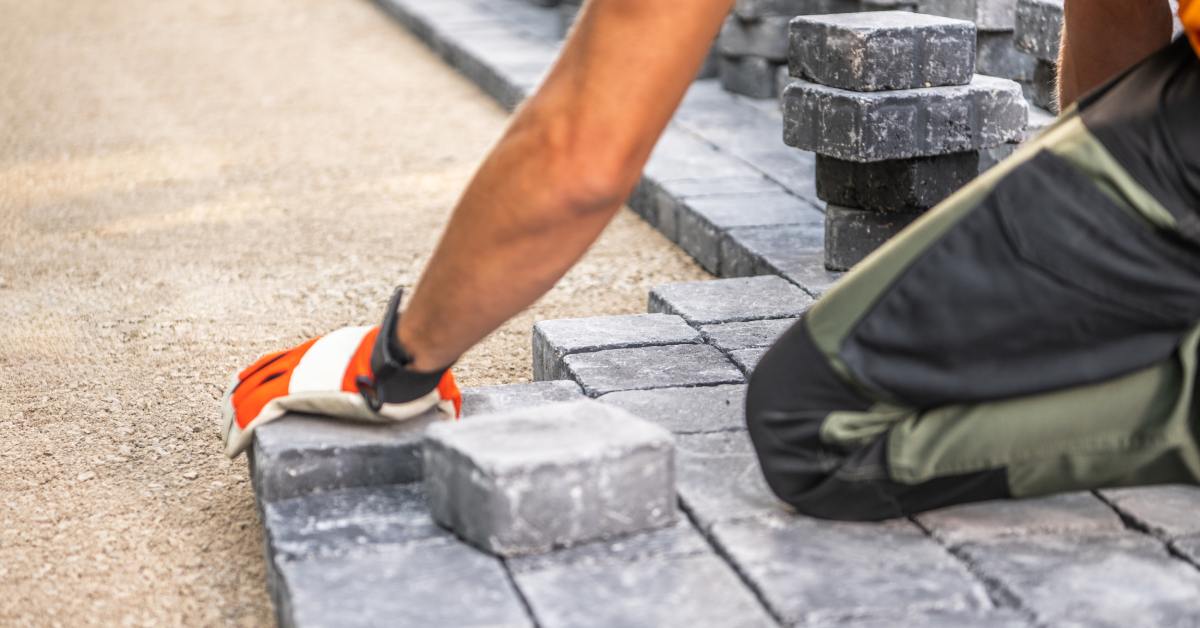 A worker laying brick pavers on a dirt surface that is leveled and cleared off.