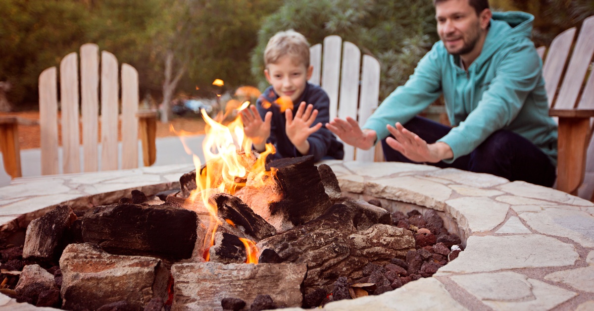 A father and his son sit in wooden Adirondack chairs next to a lit fire pit. They are warming up their hands.