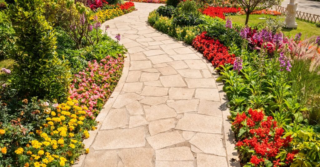 A curved flagstone pathway lined with vibrant flower beds featuring red, yellow, pink, and purple blooms in a garden.