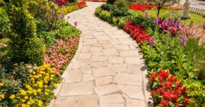 A curved flagstone pathway lined with vibrant flower beds featuring red, yellow, pink, and purple blooms in a garden.