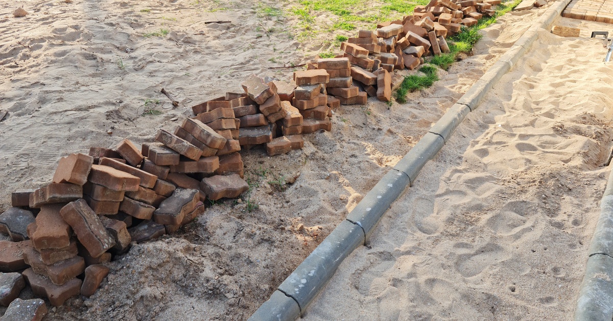 Piles of brown bricks lie on sandy ground beside a partially built pathway edged with concrete borders in bright sunlight.