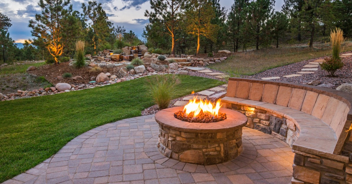 A brick outdoor fire pit overlooks a large backyard with trees and landscaping. A brick bench is next to the pit.
