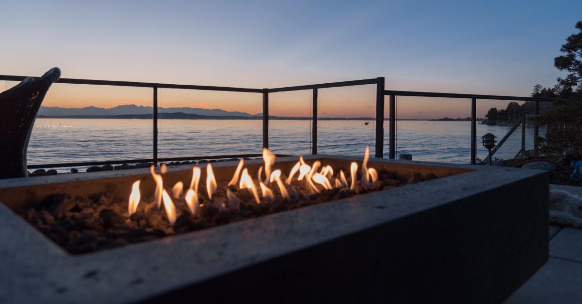 A close-up view shows a rectangular fire pit sitting on a patio that overlooks a body of water during the sunset.