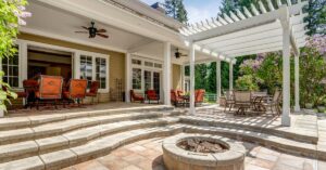 A fire pit sits on a luxurious brick patio that features a white, wooden pergola and outdoor furniture.