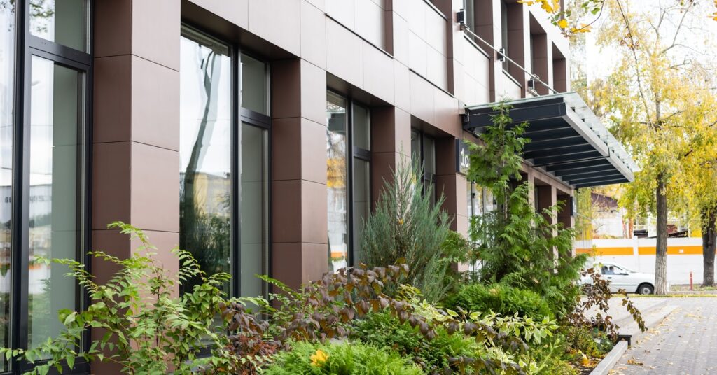 A red brick commercial building features lots of large windows. Lush, green landscaping is in front of the building.