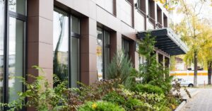 A red brick commercial building features lots of large windows. Lush, green landscaping is in front of the building.