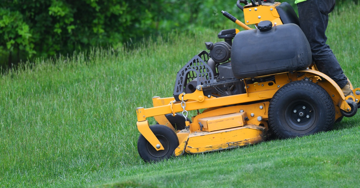 A person wearing jeans and a bright yellow shirt uses a yellow industrial lawn mower to mow a grassy area.