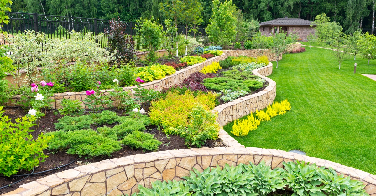 A beautifully landscaped backyard features retaining walls with elevated gardens, green grass, and trees.