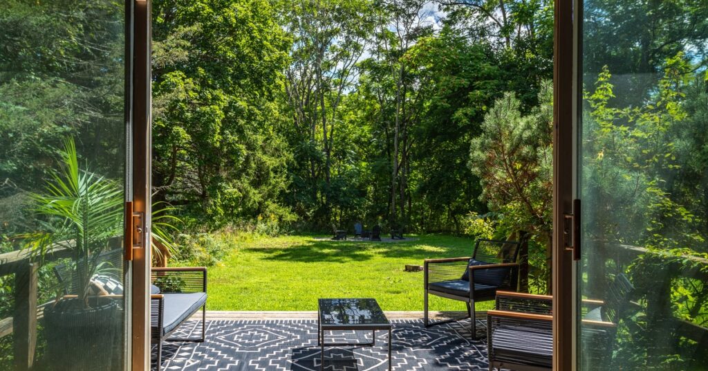 Large, glass sliding doors open to reveal a patio with a rug, outdoor furniture, and lots of greenery.