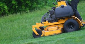 A person wearing black pants and a neon yellow shirt is using a yellow standing lawn mower to mow grass.