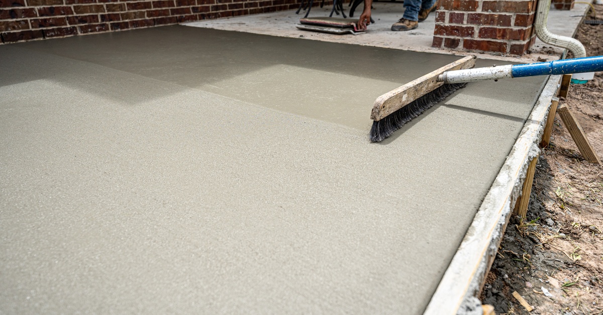 A person is pushing a broom across wet concrete, leaving behind a bumpy look. Another person is in the background.