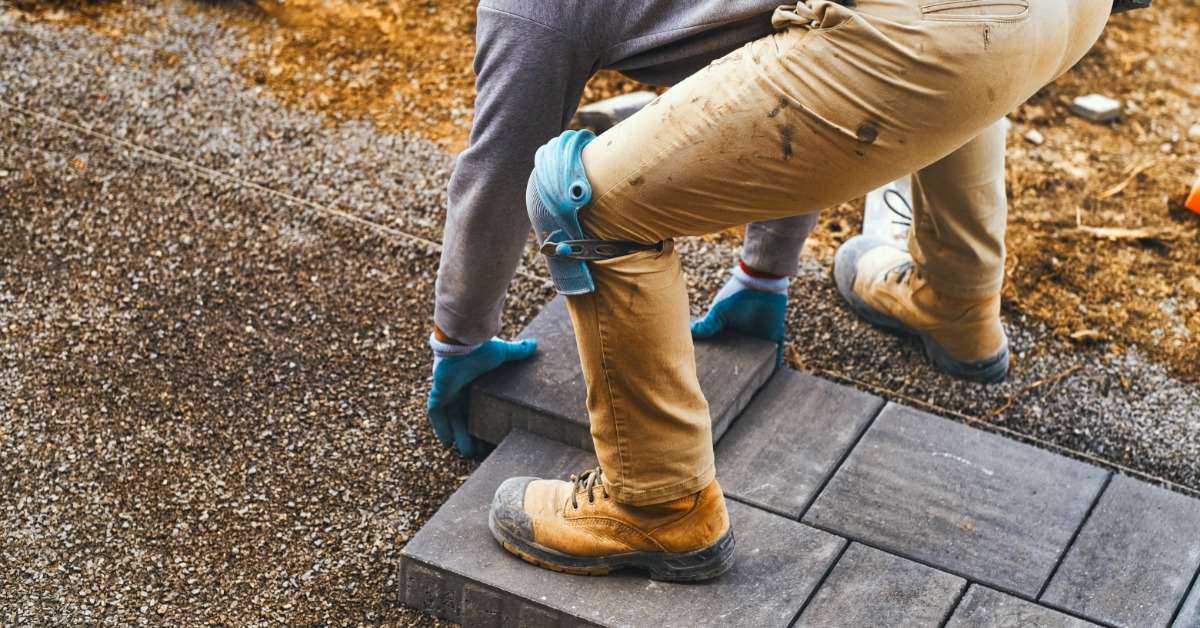 A person wearing work gloves, tan pants, and a gray long-sleeve shirt is laying dark-gray pavers on a pebble path.