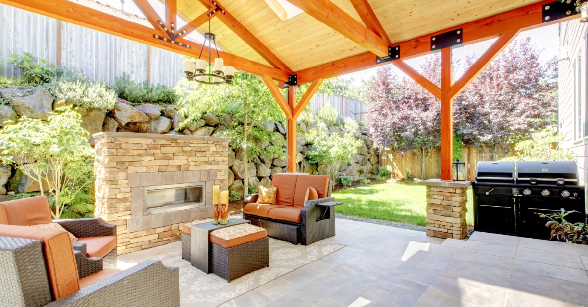 A concrete patio is covered by a wooden structure and contains a grill, patio furniture, a fireplace, and a rug.