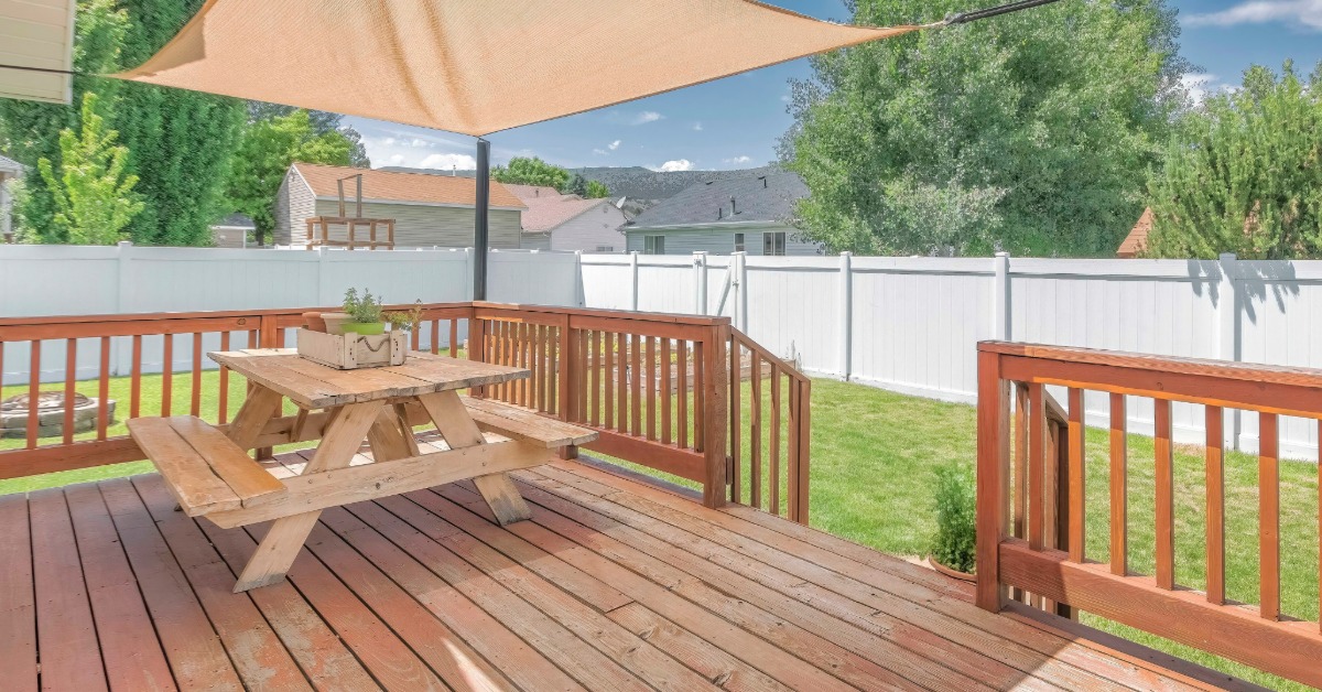 A wooden deck sits in a fenced-in backyard. A wooden picnic table sits on a deck, and a sunshade provides cover.
