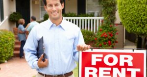 A smiling man wearing business attire tucks a clipboard under his armpit and rests a hand on a for rent sign.