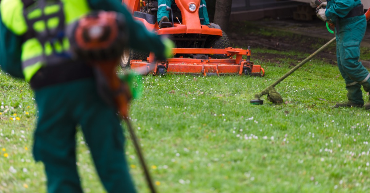 One man is mowing the grass with an orange riding lawn mower while two men use a string trimmer to trim the edges.
