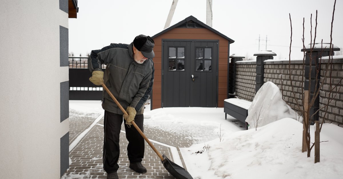 A man wearing a jacket and a winter hat is shoveling the snow on a walkway in the fenced-in backyard.